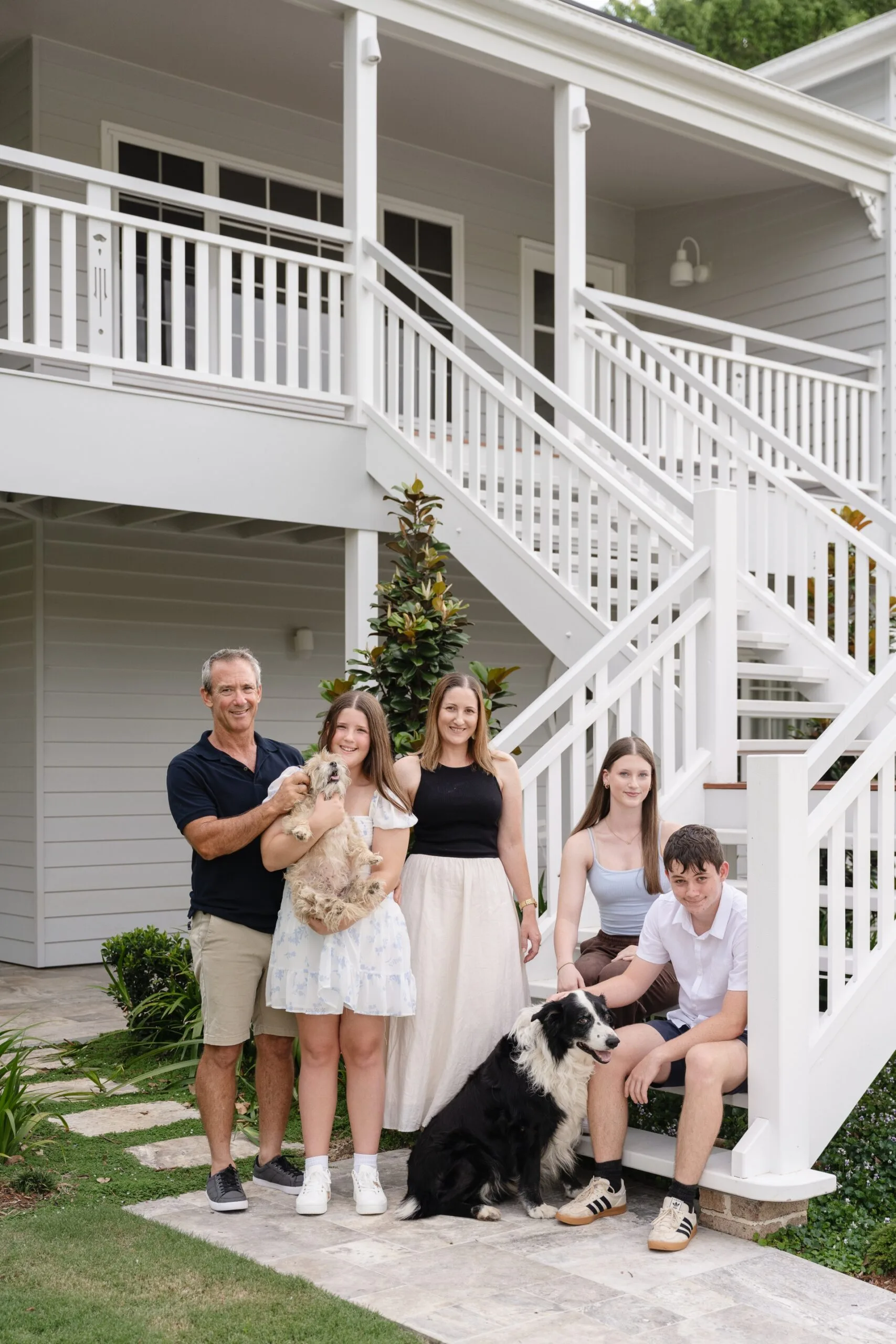 Front house clear mountain family members Nathan, Macey (holding Tess, the Maltese cross), Belinda, Ellie, Tyler and Charlie, the Border Collie, gather at the bottom of the entry staircase.