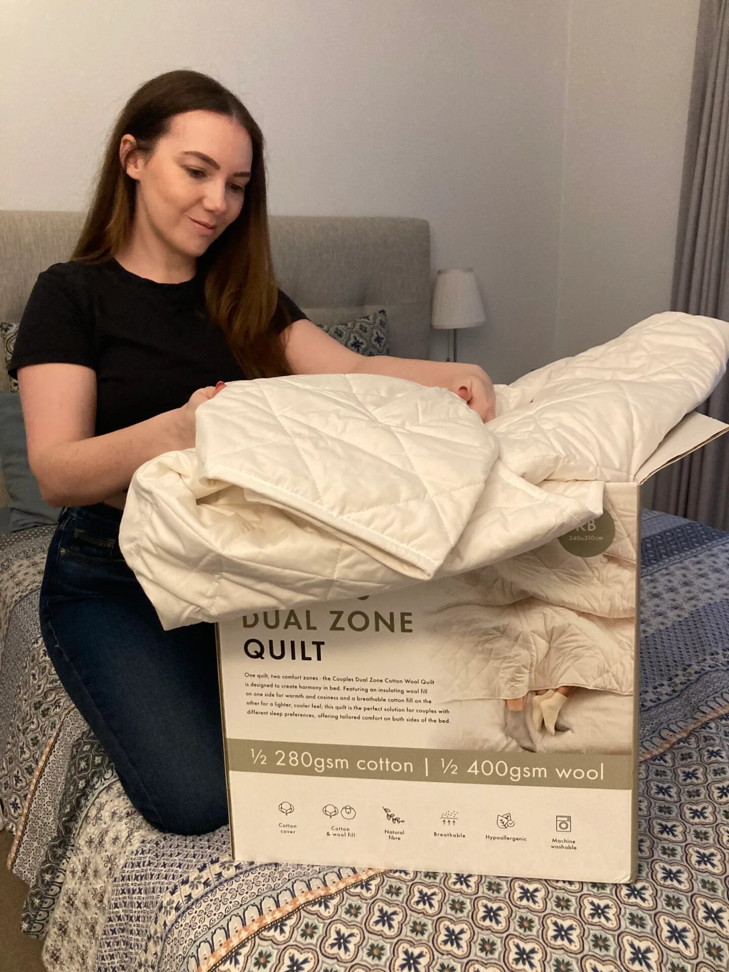 A woman with brown hair kneels on a bed, opening a cardboard box and removing the white quilt inside.