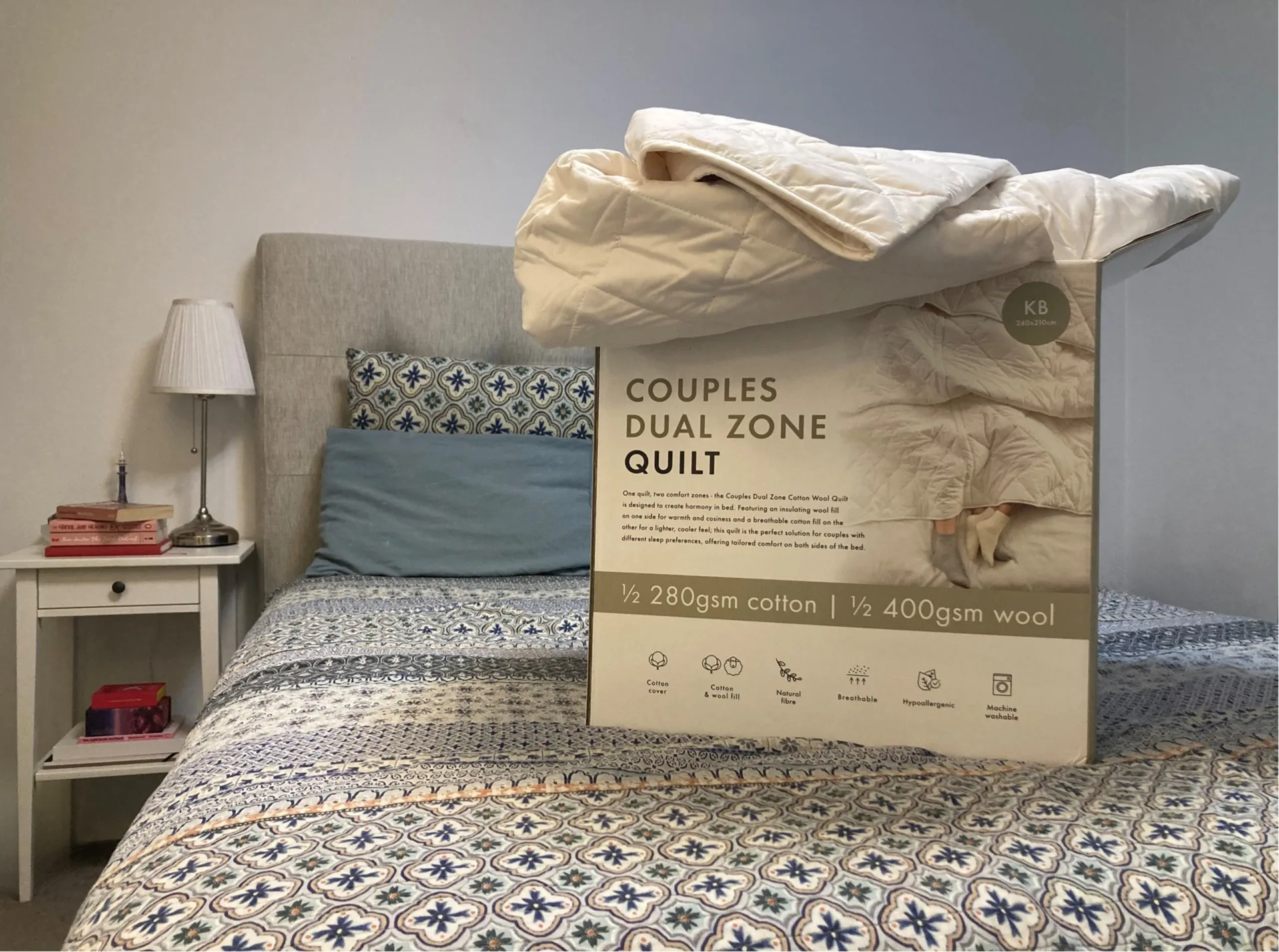 A cardboard box with a quilt coming out of the top is positioned on a blue patterned quilt, on a bed. A bedside table has a lamp and two stacks of books on it.
