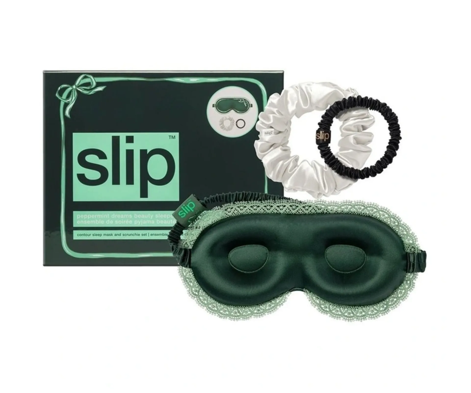 A dark green contoured silk sleep mask with a lighter green lace trim, white scrunchie, and black, smaller scrunchie, positioned on top of a gift box labelled 'Slip'