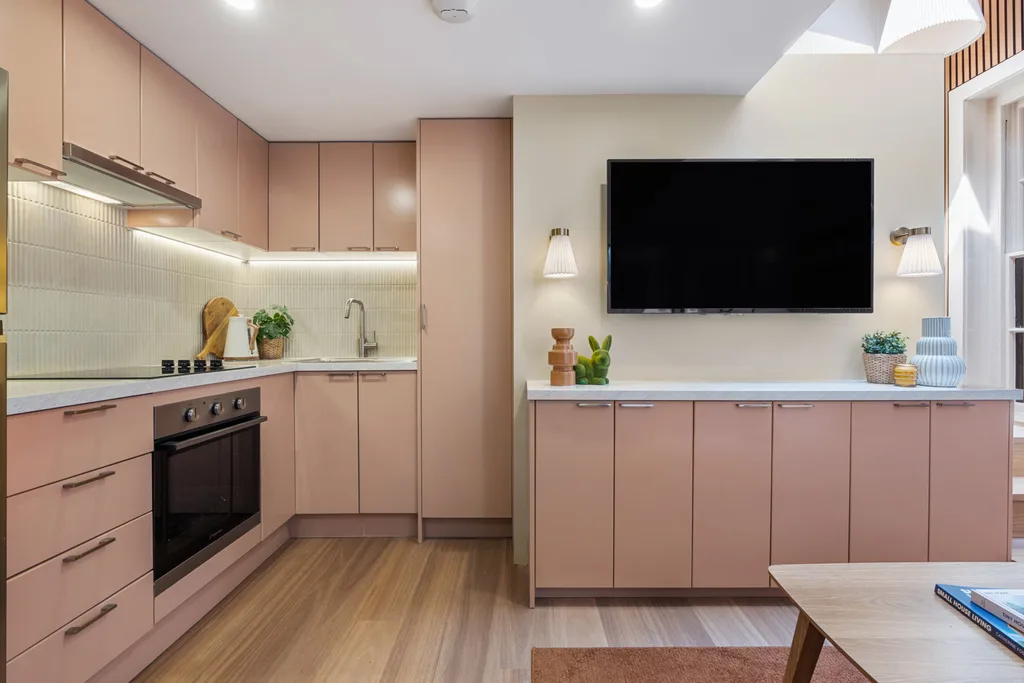 An apartment kitchen with pink cabinetry, white benchtops and white kit kat tiled splashback. Hardware is chrome. To the right of the kitchen, the cabinetry continues underneath a wall-mounted TV.