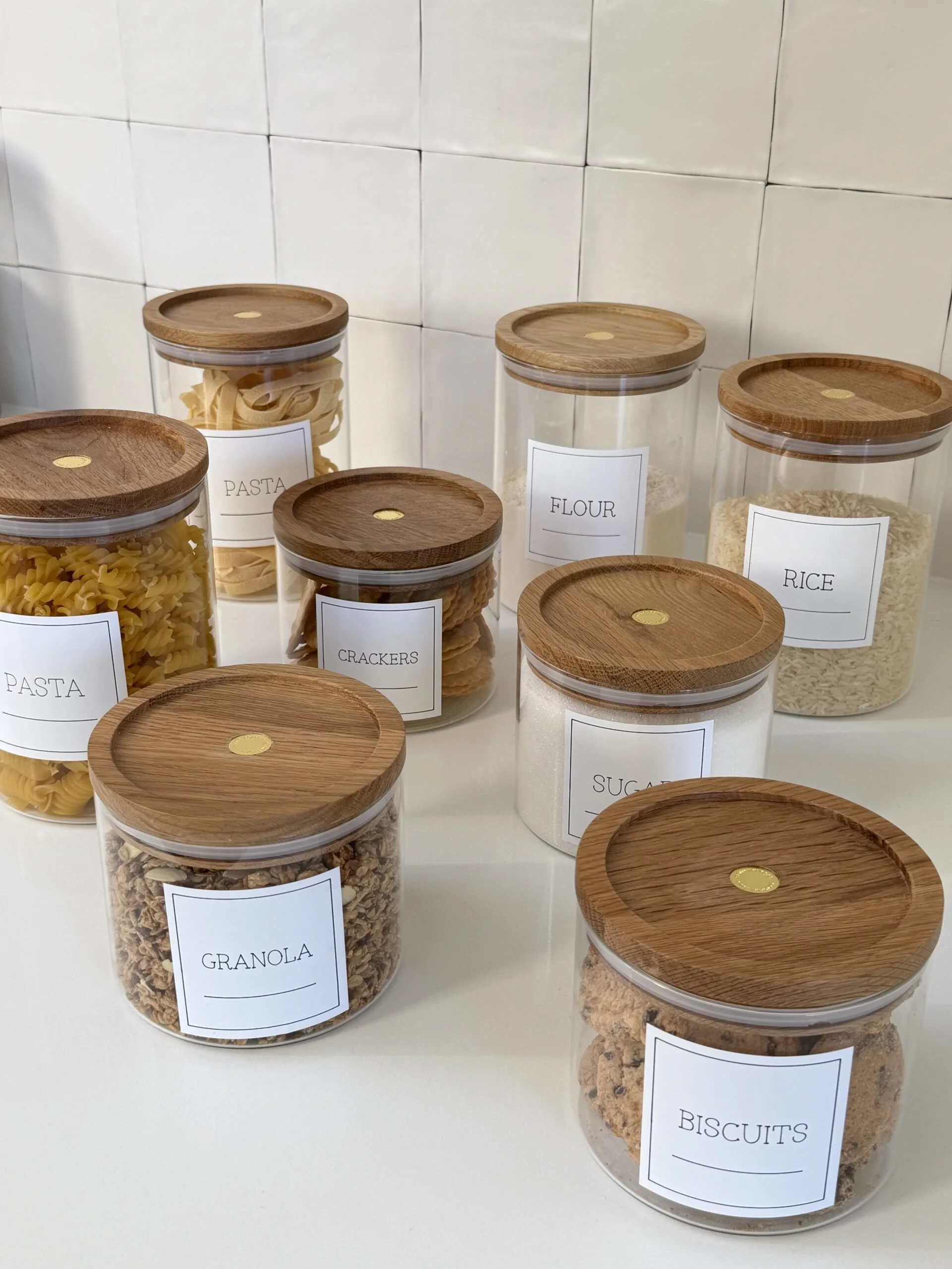 Clear jars with wooden lids labeled with pasta, crackers, flour, rice, granola, sugar, and biscuits on a white counter.