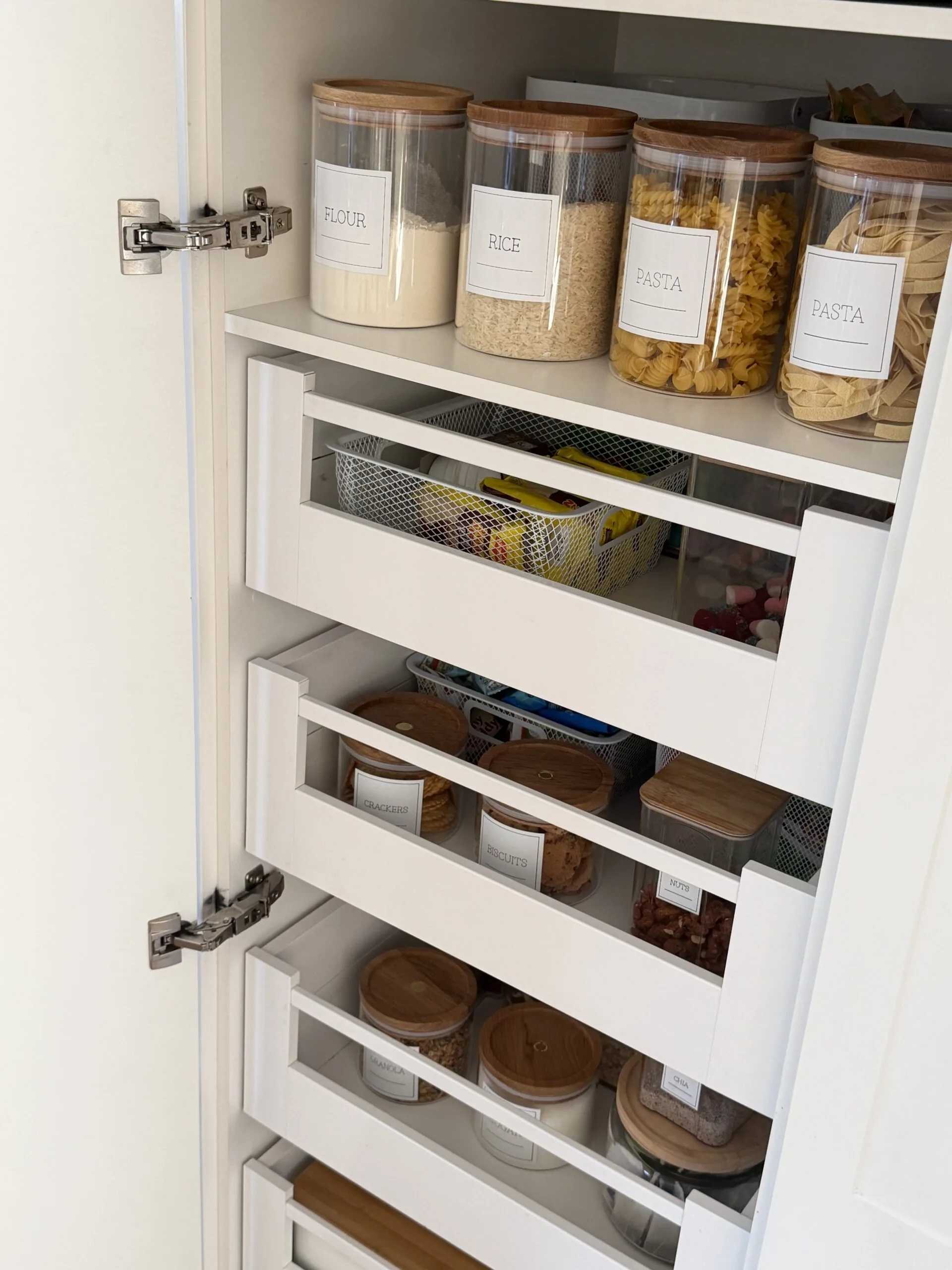 A pantry with labeled jars containing flour, rice, pasta, crackers, biscuits, and nuts neatly organized on white shelves.