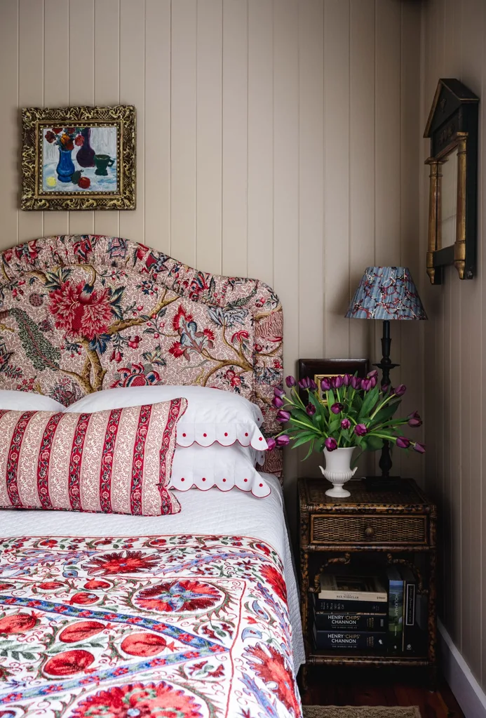 Cosy and charming bedroom with pink, blue and red floral headboard and bedspread, side table with tulips, books, and a lamp, art on beige VJ panelled wall.
