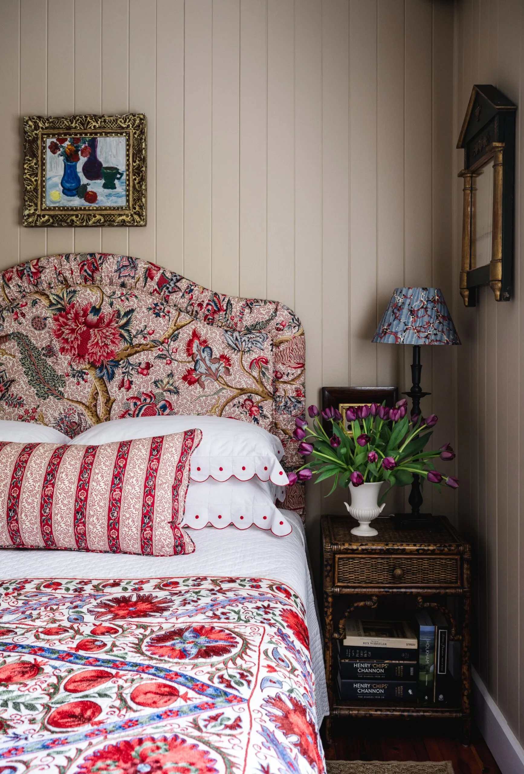 Cosy and charming bedroom with pink, blue and red floral headboard and bedspread, side table with tulips, books, and a lamp, art on beige VJ panelled wall.