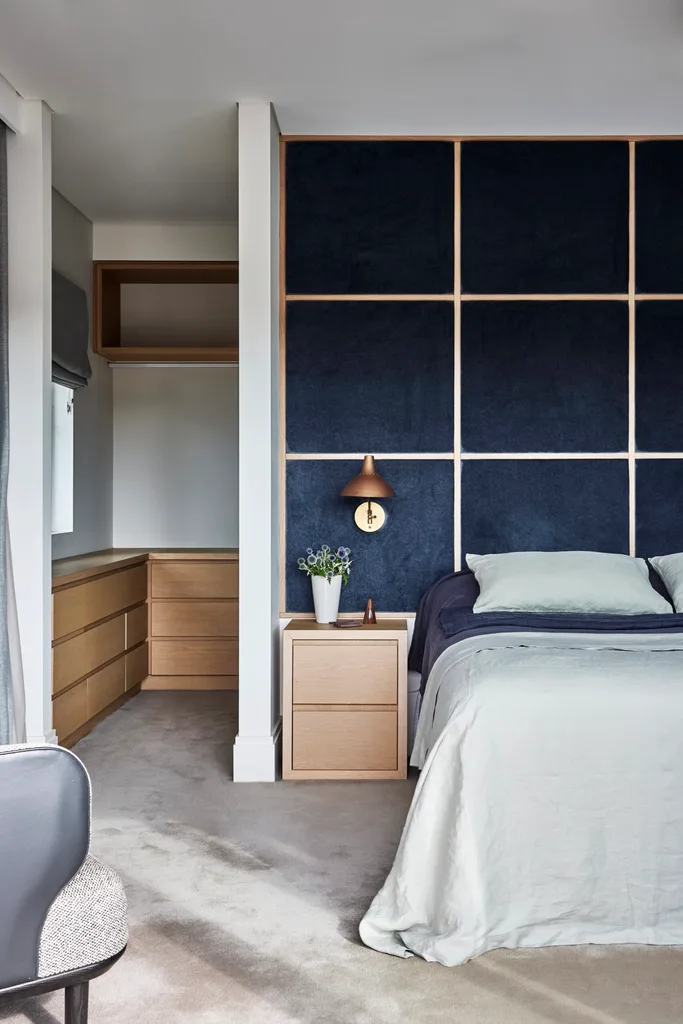 Bedroom with a navy accent wall, wooden nightstand, copper lamp, and a doorway leading to a closet with wooden drawers. The bed has a mix of navy and light blue bedding on it.
