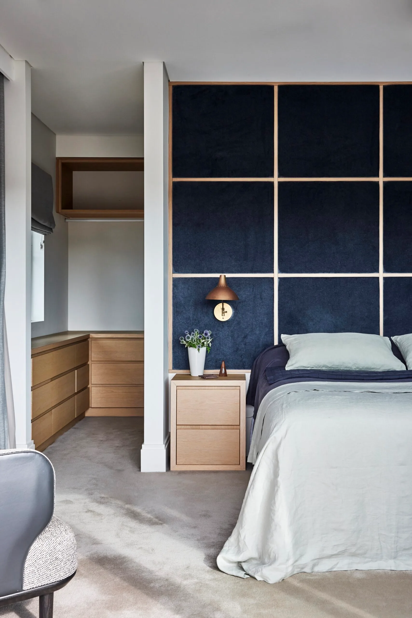Bedroom with a navy accent wall, wooden nightstand, copper lamp, and a doorway leading to a closet with wooden drawers. The bed has a mix of navy and light blue bedding on it.