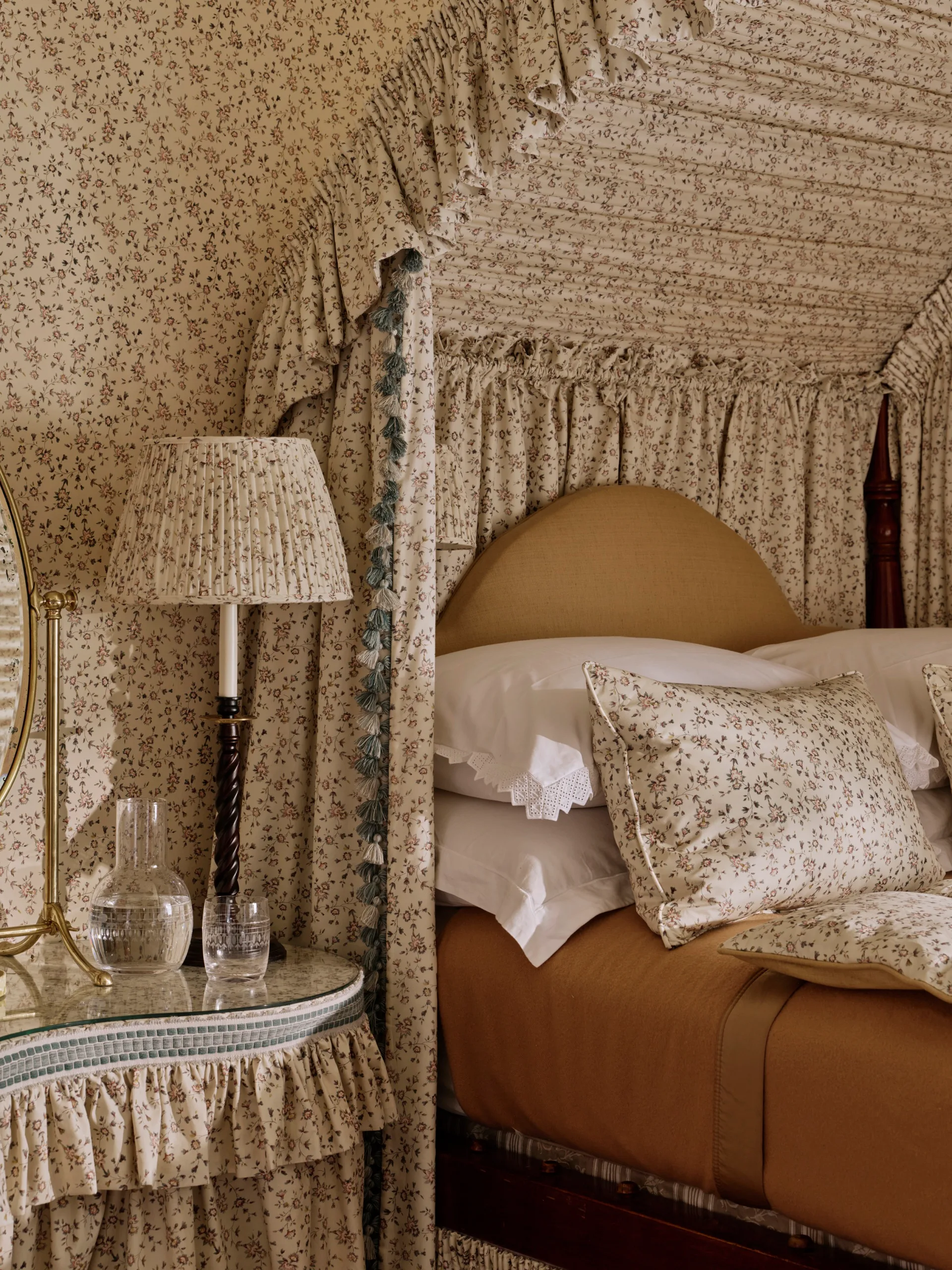 Floral-patterned bedroom with canopy bed, matching lampshade, and a decorated side table with carafe and glass.