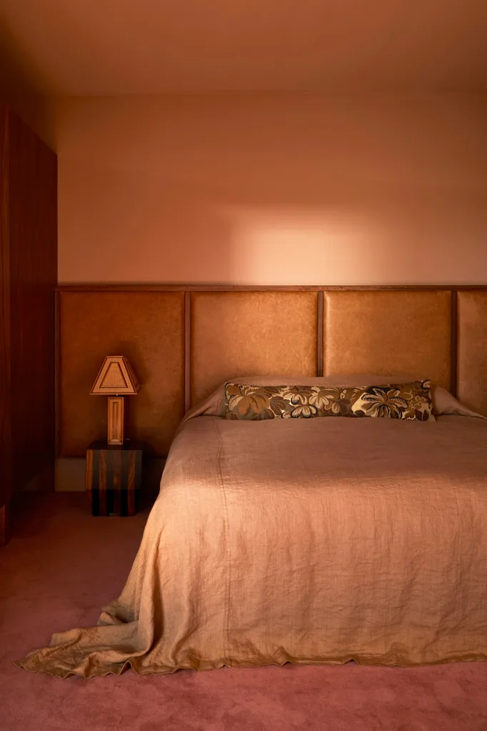 A cosy colour drenched bedroom with a dusty pink bedspread, floral pillow, and a wooden lamp on a nightstand against a soft pink wall. The carpet is a deep pink.