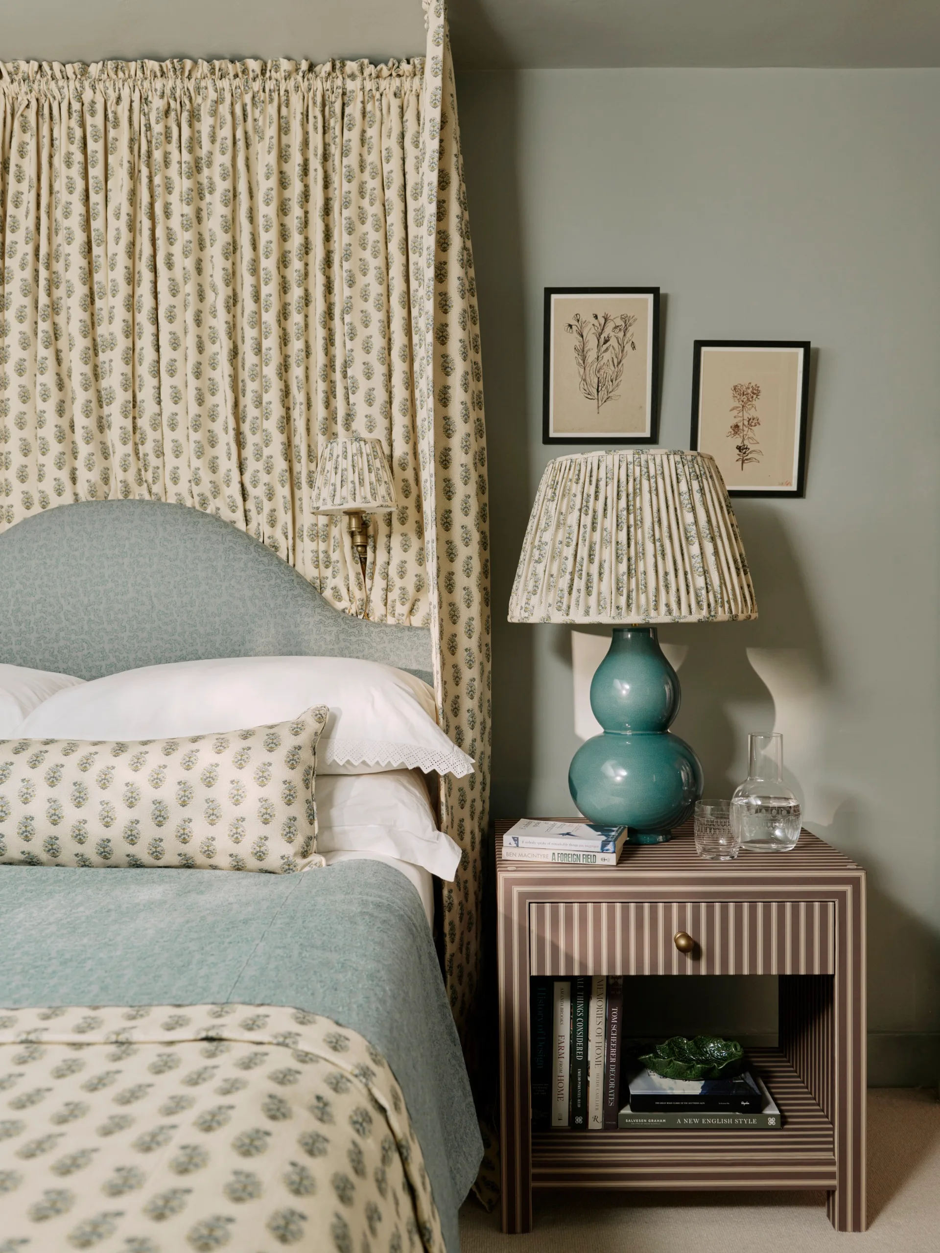 Bedroom with patterned drapes, matching lampshades and bedding. A striped nightstand is beside the blue upholstered headboard, decorated with books, a blue lamp, and botanical wall art.