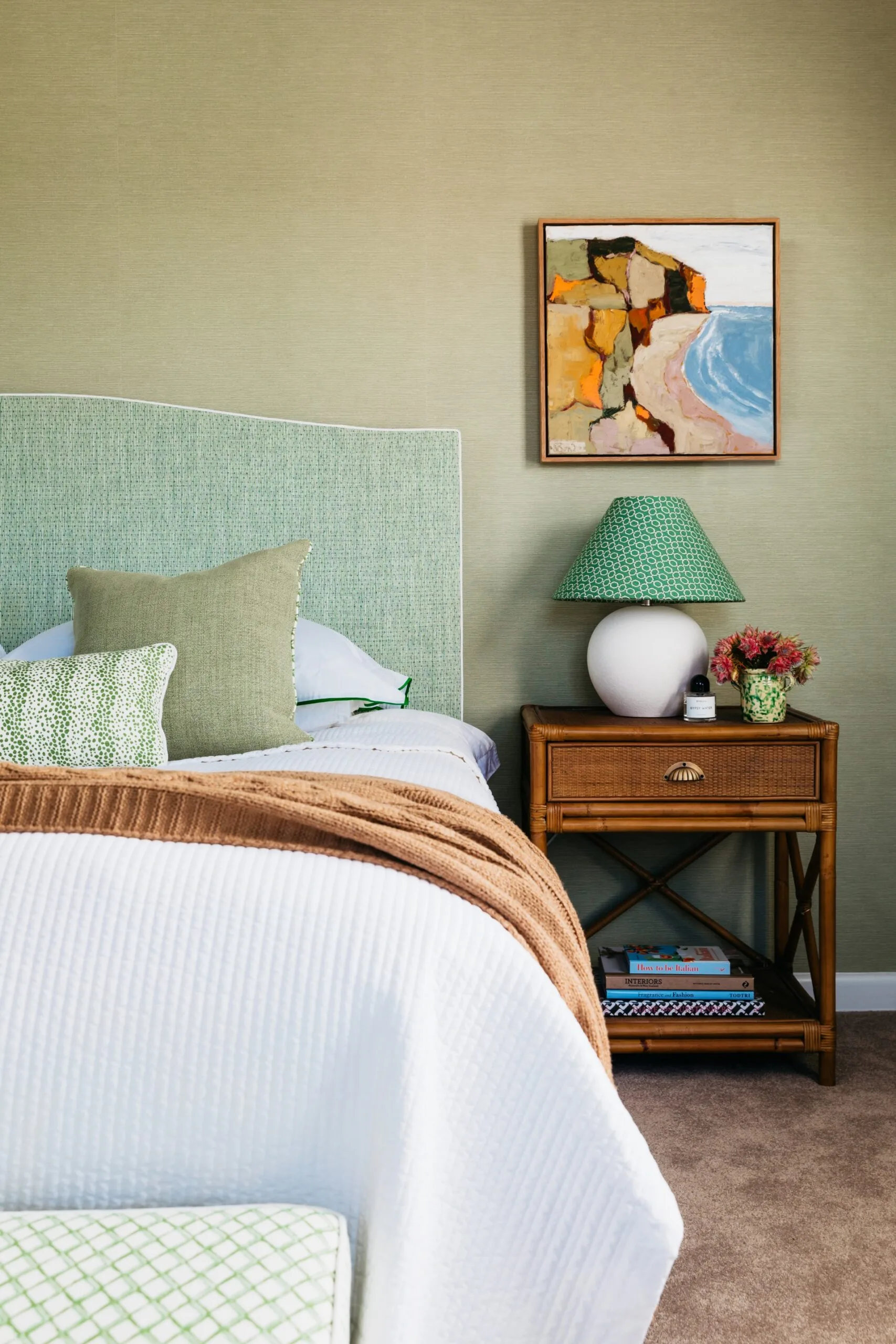 Bed with green headboard, pillows, and a tan throw. Nightstand with lamp, flowers, and art on the wall.