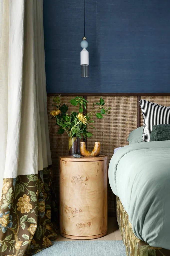 Colourful bedroom with floral curtain, wicker headboard, round wood nightstand, and a modern pendant light.