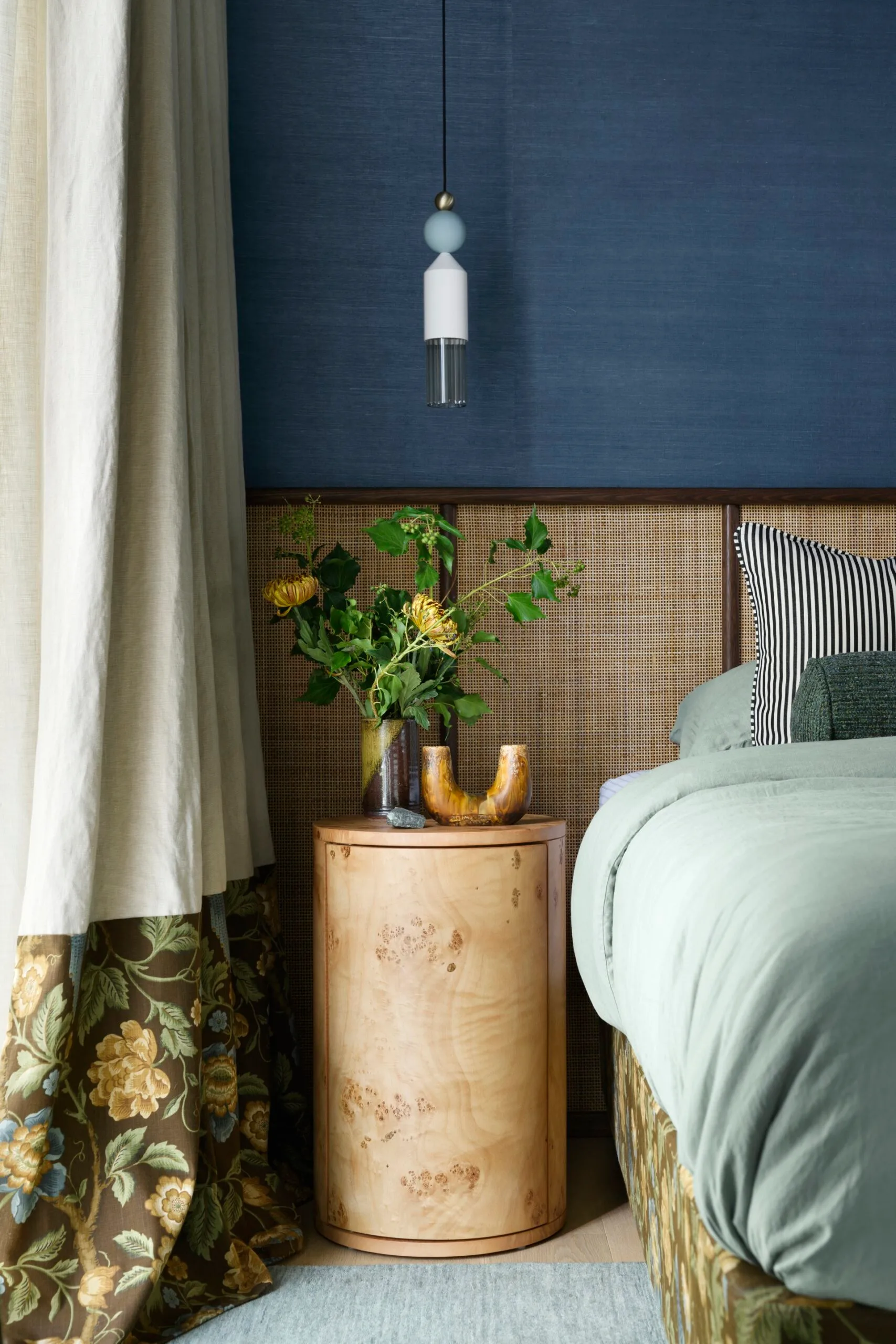 Colourful bedroom with floral curtain, wicker headboard, round wood nightstand, and a modern pendant light.