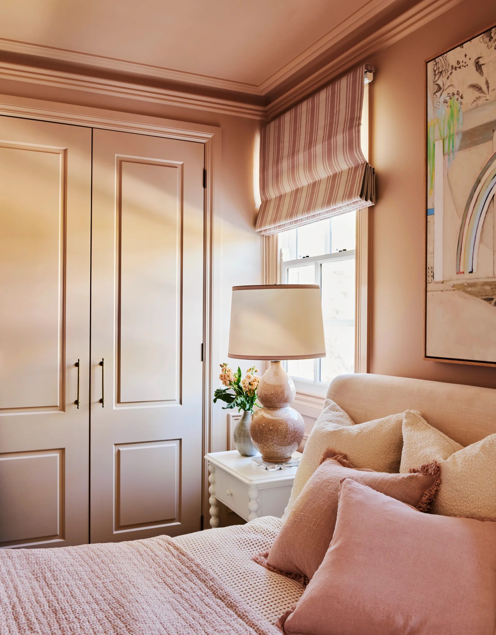 Bedroom with pink walls, a pink bed with pillows, a side table with a lamp, and a window with striped blinds. The headboard, blankets and pillows on the bed all feature different shades of pink, from creamier softer tones to darker ones.