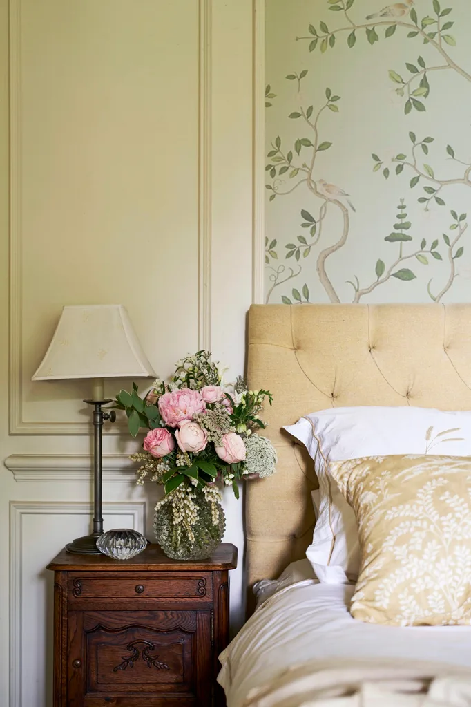 Cosy bedroom with a gentle and calming bedroom colour scheme. Floral wallpaper and panelled warm white walls are behind a soft yellow tufted headboard. Beside the bed is a wooden nightstand, lamp, and vase of pink flowers.