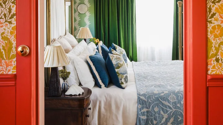Eclectic and bright bedroom with green curtains, floral wallpaper. In the foreground is the doorway leading to the bedroom. The architraves and surrounding wainscoting is a vibrant red colour. Above is floral wallpaper in autumnal colours. Inside the bedroom, the bed features blue and white patterned bedding.