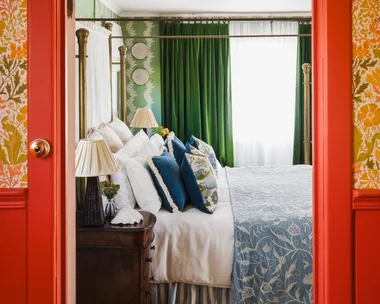 Eclectic and bright bedroom with green curtains, floral wallpaper. In the foreground is the doorway leading to the bedroom. The architraves and surrounding wainscoting is a vibrant red colour. Above is floral wallpaper in autumnal colours. Inside the bedroom, the bed features blue and white patterned bedding.