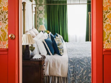 Eclectic and bright bedroom with green curtains, floral wallpaper. In the foreground is the doorway leading to the bedroom. The architraves and surrounding wainscoting is a vibrant red colour. Above is floral wallpaper in autumnal colours. Inside the bedroom, the bed features blue and white patterned bedding.
