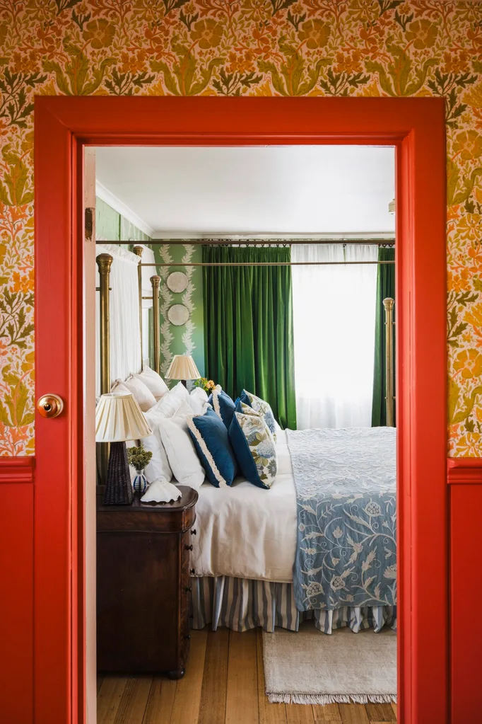 Eclectic and bright bedroom with green curtains, floral wallpaper. In the foreground is the doorway leading to the bedroom. The architraves and surrounding wainscoting is a vibrant red colour. Above is floral wallpaper in autumnal colours. Inside the bedroom, the bed features blue and white patterned bedding.