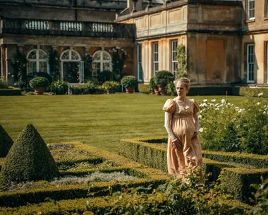 A still from period drama Sanditon. A woman walks between two low hedges in the grounds of a grand Georgian estate.