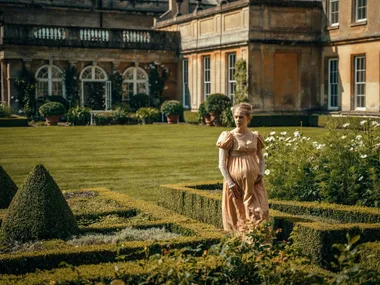 A still from period drama Sanditon. A woman walks between two low hedges in the grounds of a grand Georgian estate.