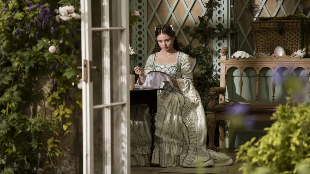 A still from Queen Charlotte: A Bridgerton Story. A woman in a grand Regency-ish era dress (not particularly historically accurate) sits at a desk embroidering with a sewing hoop. Flowers surround her.
