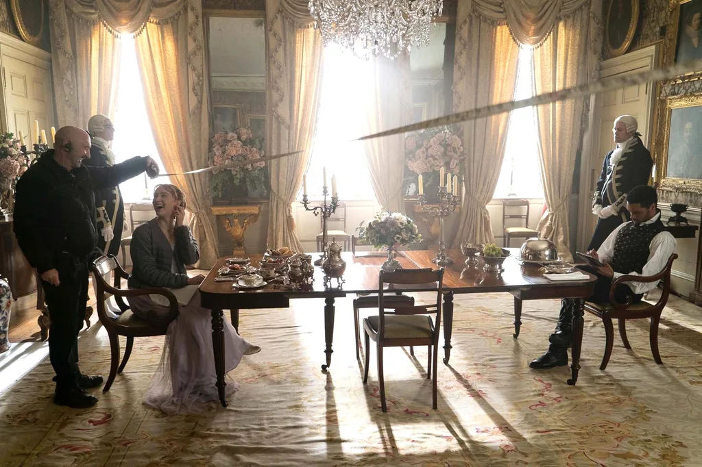 A photo of a film set (Bridgerton, season one). Two actors sit at a grand dining table, with two butlers nearby. A rug covers the ground while a grand chandelier is positioned above. A film crew holds a measuring tape near one of the actors. They are both laughing.