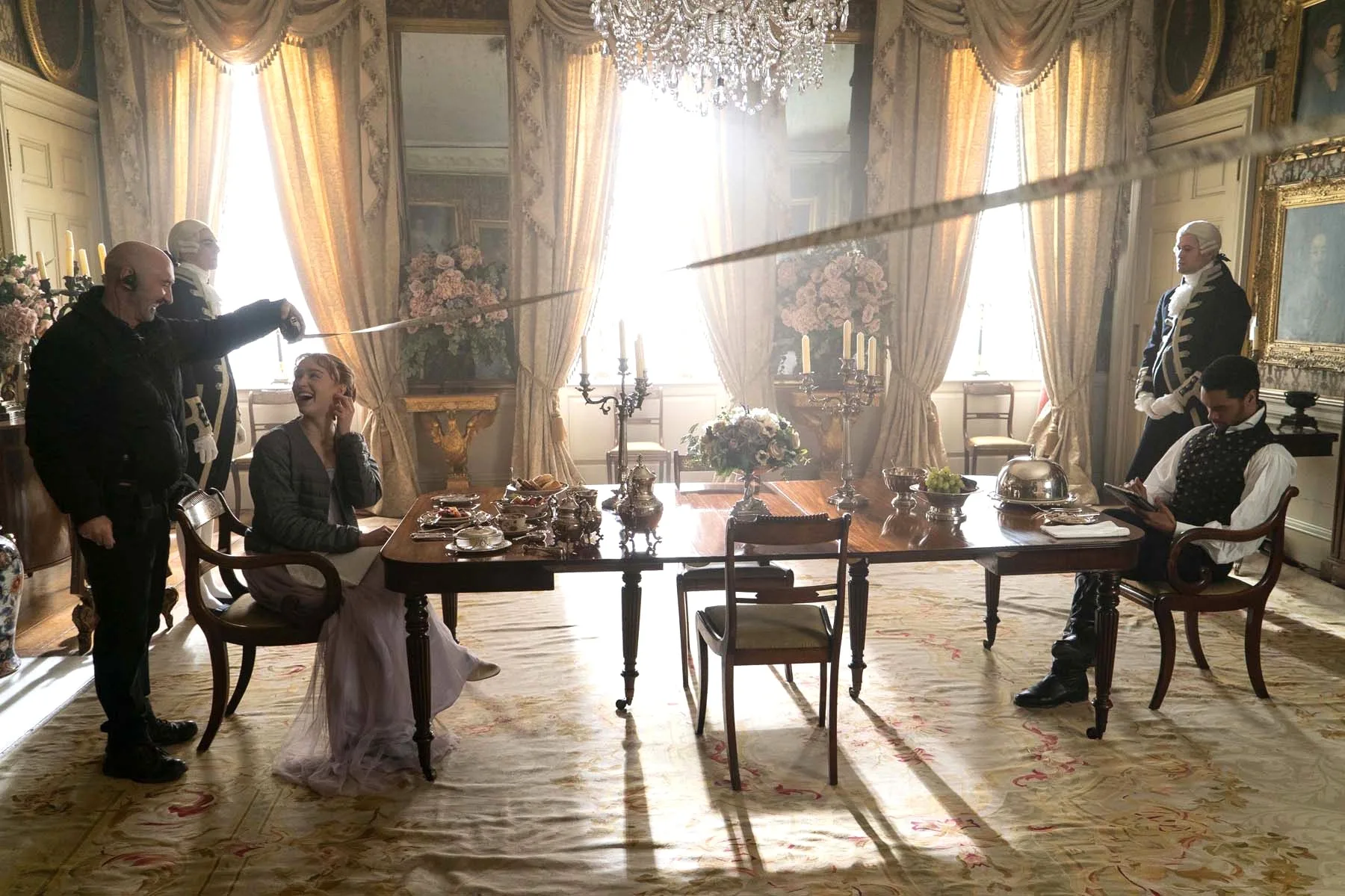 A photo of a film set (Bridgerton, season one). Two actors sit at a grand dining table, with two butlers nearby. A rug covers the ground while a grand chandelier is positioned above. A film crew holds a measuring tape near one of the actors. They are both laughing.