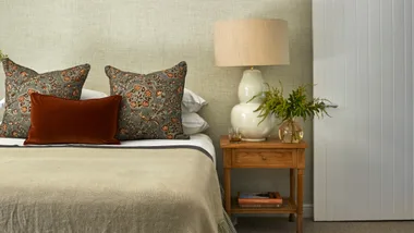 Bed with floral cushions, a brown velvet pillow, a wooden nightstand, table lamp, green plant, and books.