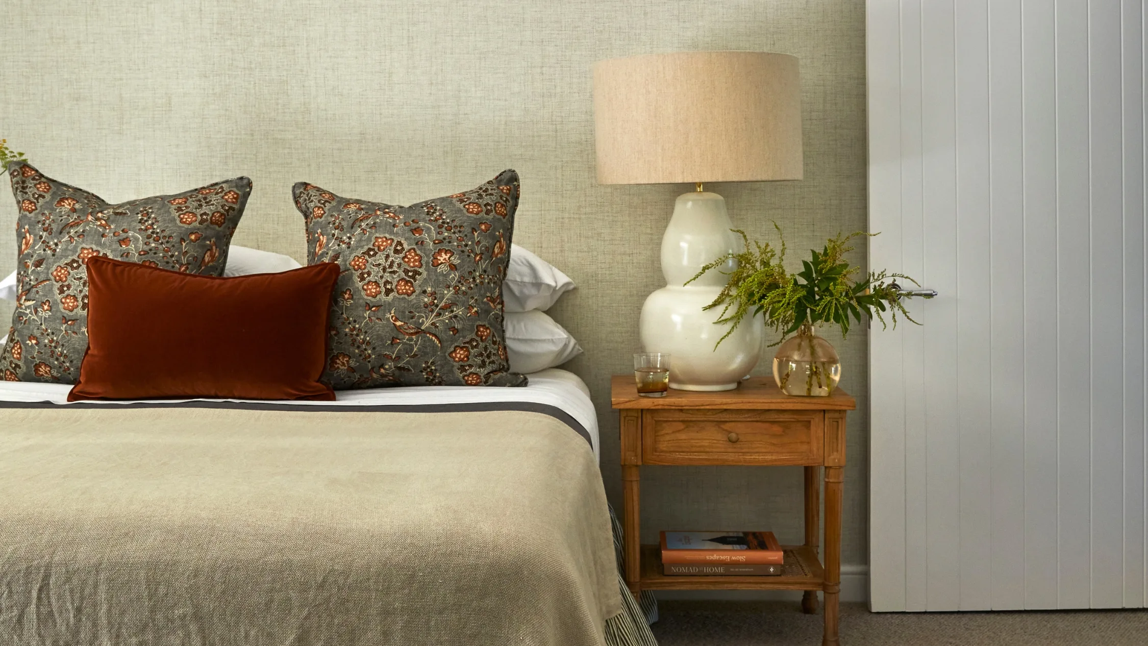 Bed with floral cushions, a brown velvet pillow, a wooden nightstand, table lamp, green plant, and books.