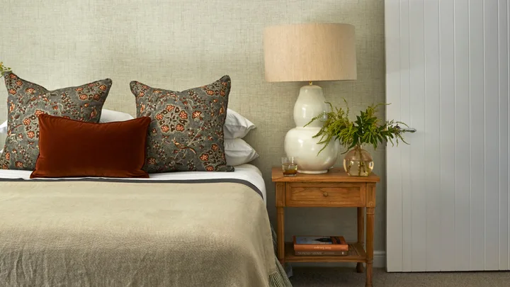 Bed with floral cushions, a brown velvet pillow, a wooden nightstand, table lamp, green plant, and books.