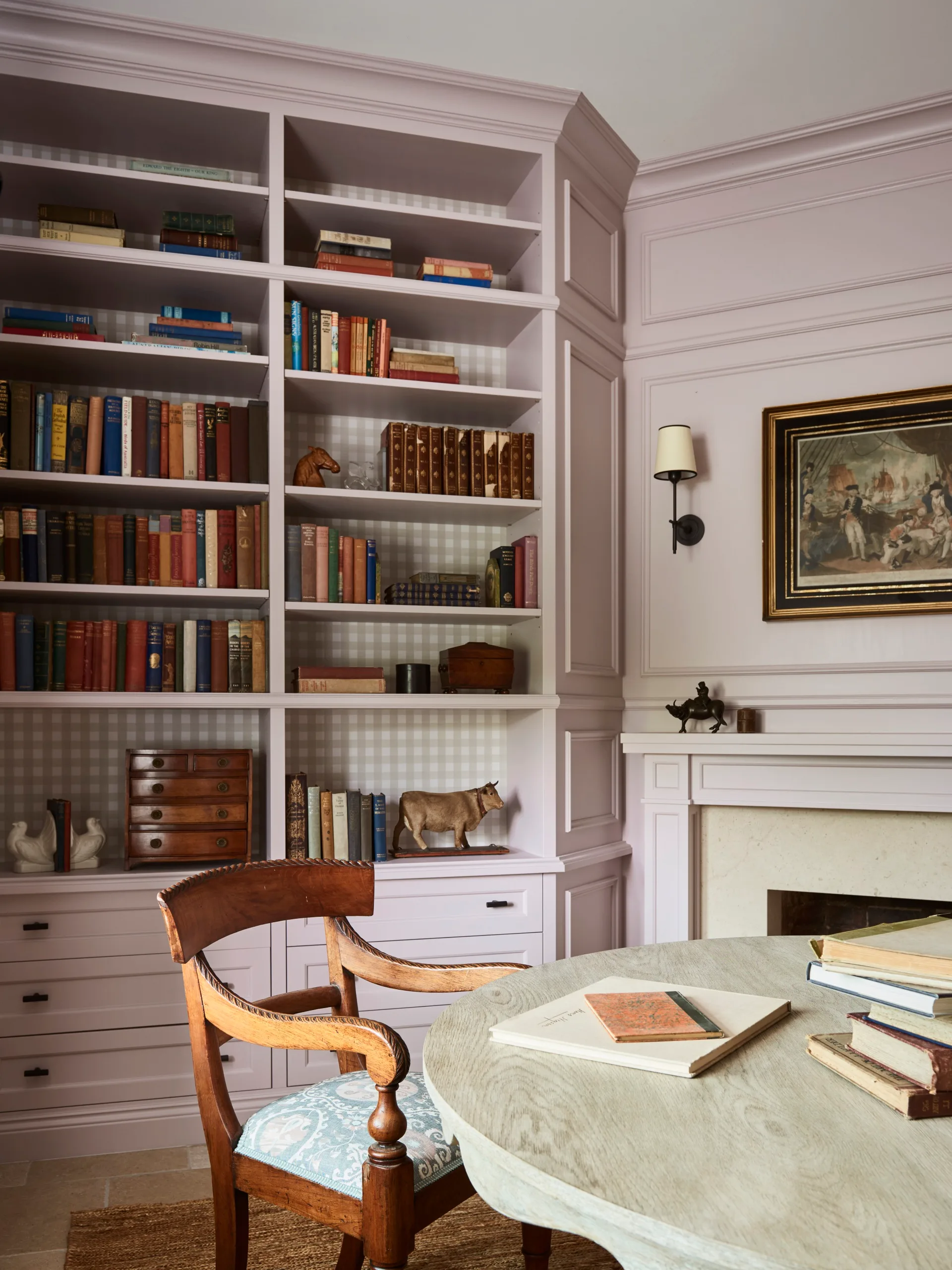 Cosy home library with pastel lilac built-in bookshelves, books, wooden chair, and a round table, featuring decorative items and painting above a fireplace.
