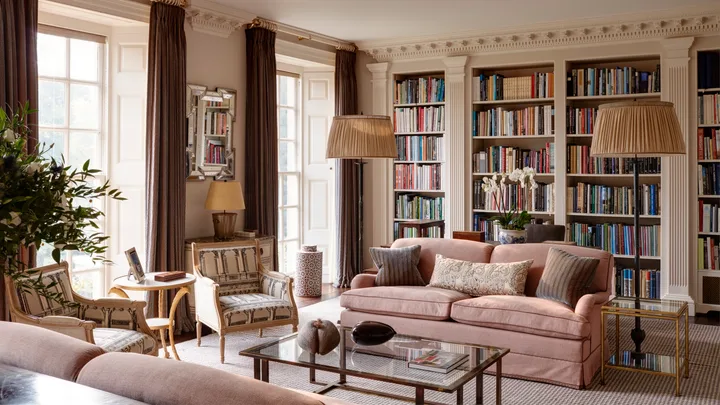 Luxurious home library room with a pink sofa, bookshelves, ornate decorative columns between the built-in bookshelves, armchairs, and large windows with drapes. Two floor lamps are positioned on either side of the pink sofa and a glass coffee table is positioned in front.