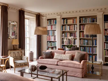 Luxurious home library room with a pink sofa, bookshelves, ornate decorative columns between the built-in bookshelves, armchairs, and large windows with drapes. Two floor lamps are positioned on either side of the pink sofa and a glass coffee table is positioned in front.