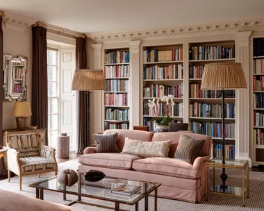 Luxurious home library room with a pink sofa, bookshelves, ornate decorative columns between the built-in bookshelves, armchairs, and large windows with drapes. Two floor lamps are positioned on either side of the pink sofa and a glass coffee table is positioned in front.