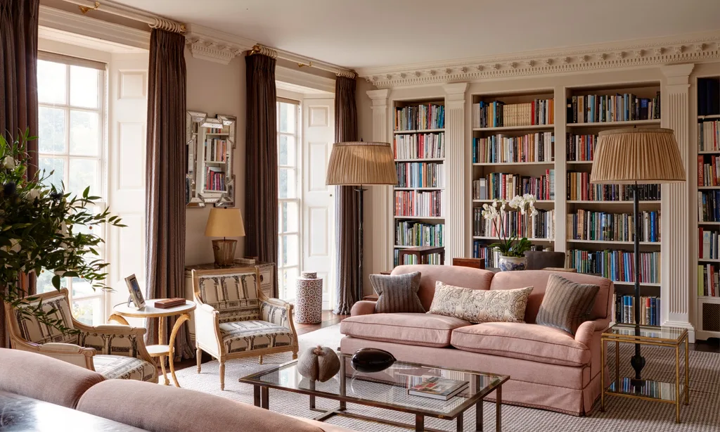 Luxurious home library room with a pink sofa, bookshelves, ornate decorative columns between the built-in bookshelves, armchairs, and large windows with drapes. Two floor lamps are positioned on either side of the pink sofa and a glass coffee table is positioned in front.