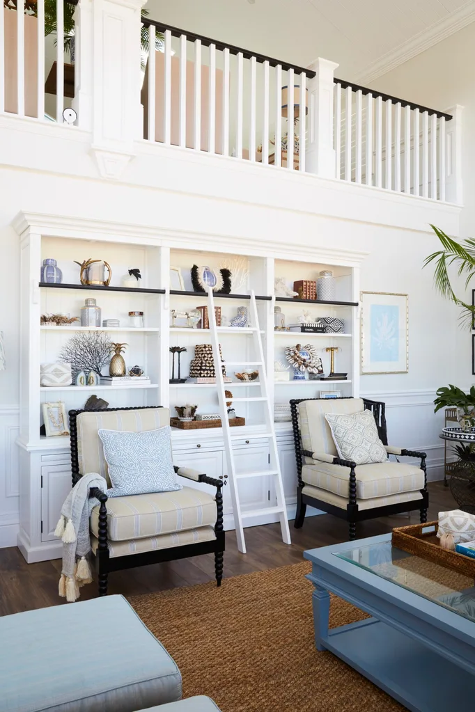 Elegant living room or home library with white shelves, decorative objects, striped armchairs, a ladder, and potted plant. The bookshelves are covered in books and decorative objects from around the world while a white library ladder leans against the shelves.
