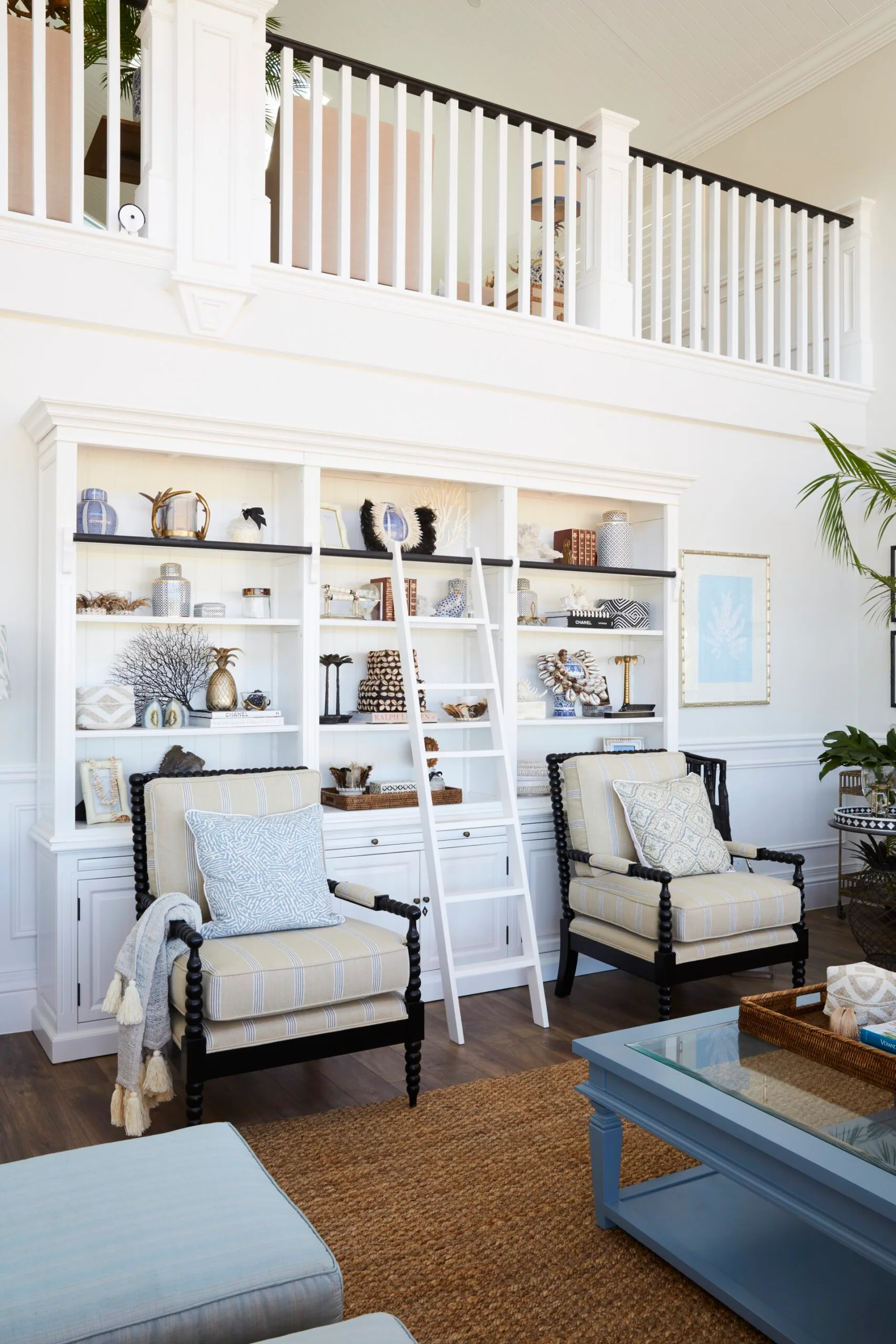 Elegant living room or home library with white shelves, decorative objects, striped armchairs, a ladder, and potted plant. The bookshelves are covered in books and decorative objects from around the world while a white library ladder leans against the shelves.