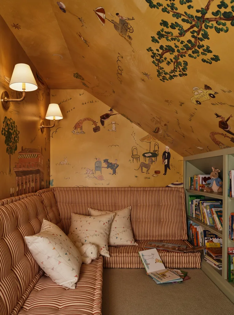 A home library or book nook in an attic for a child. Striped cushions create an upholstered seat along the wall, in an L-shape. Above this, wallpaper with a whimsical Parisian city print covers the walls and sloping ceiling. On the right hand side of the room, where the ceiling slopes down to its lowest point, green bookshelves are built in to the wall. The shelves are covered with children's books and a soft Peter Rabbit bear.