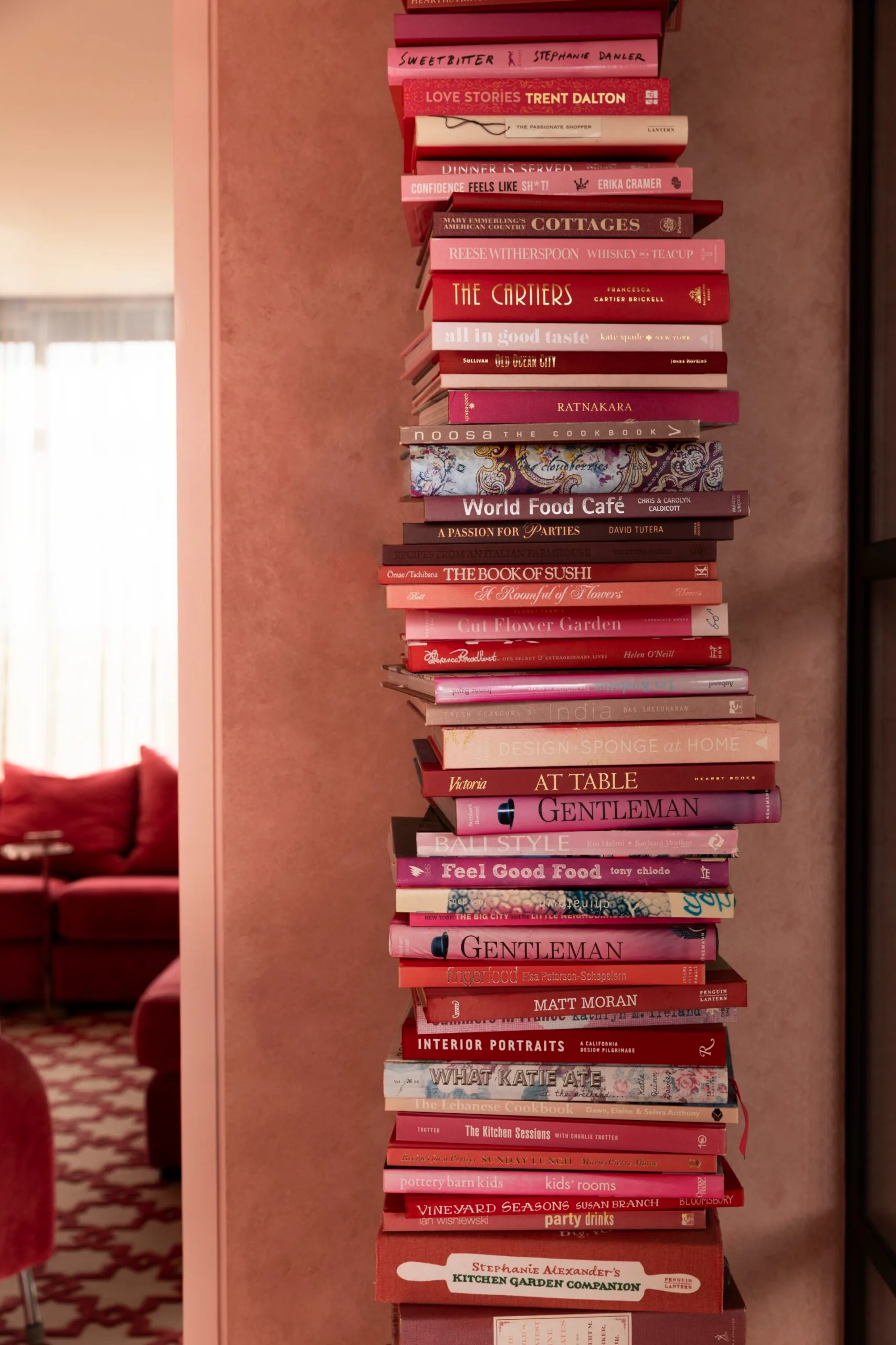 A tall stack of books in a tonal scheme. They are all varying shades of pink, from baby pink to fuschia and magenta and even red. The stack is in front of a soft pink toned wall.