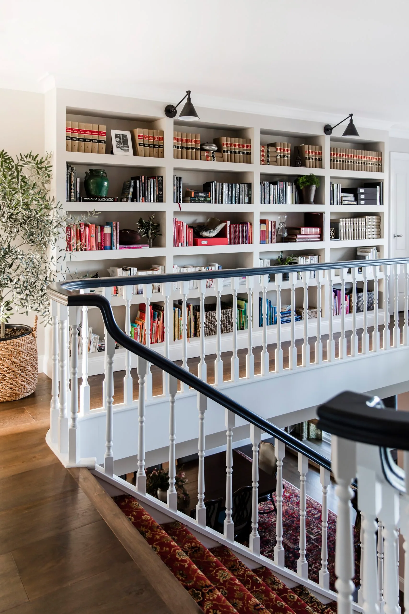 Upstairs hallway that functions as a home library with white railings forming a boundary against the timber and red carpeted stairs. A large built-in bookshelf is filled with books, two black sconces are positioned above and a potted plant is to the side in a wicker basket.