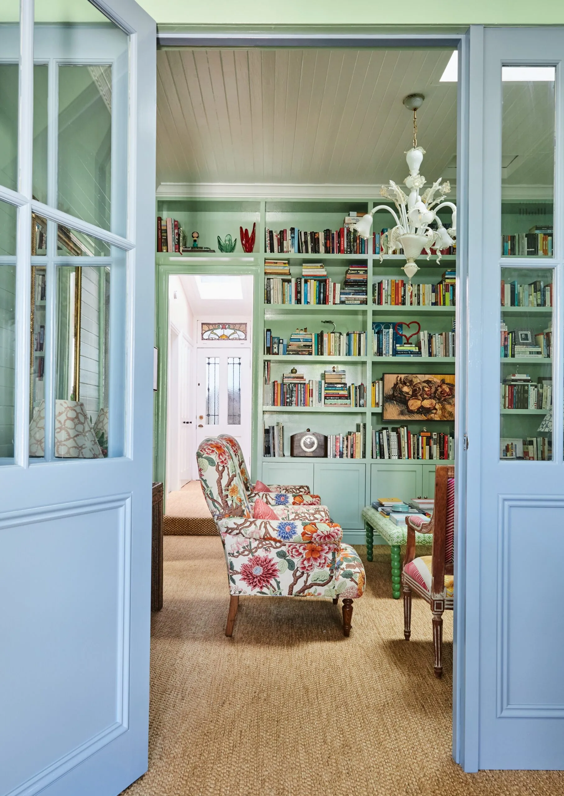 Blue doors with glass panes open to reveal a green home library with sisal carpet. Built-in bookshelves line the room decorated with books and art. A floral patterned armchair is in the middle of the room, infront of a green upholstered ottoman which also has books stacked on it.