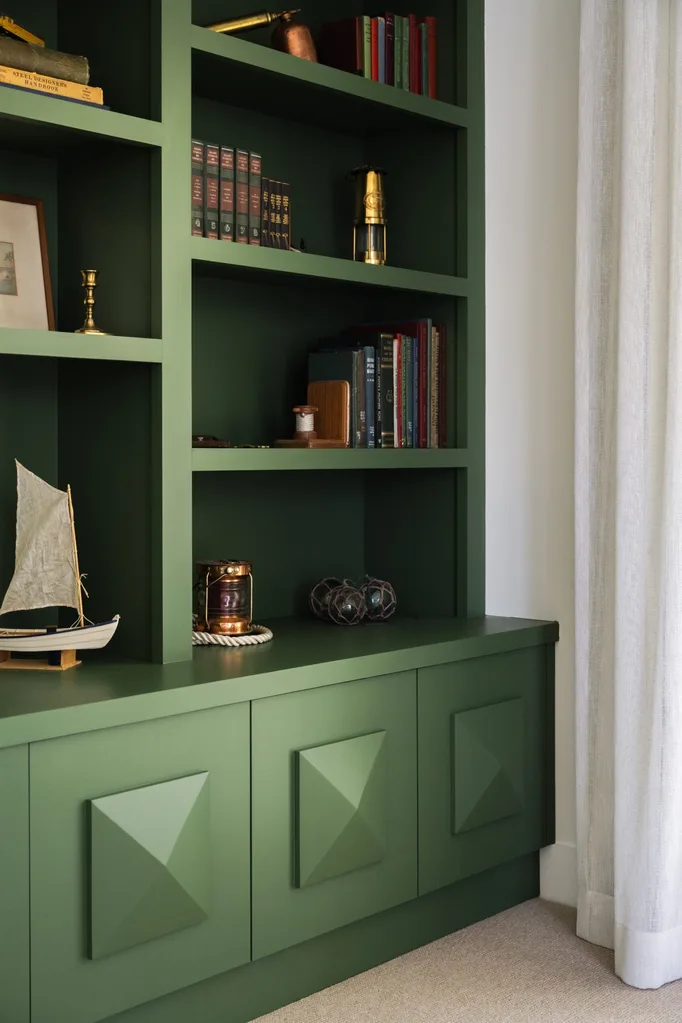Green built-in bookshelf with books, nautical decor, and brass items, featuring a small sailboat model on a lower shelf.
