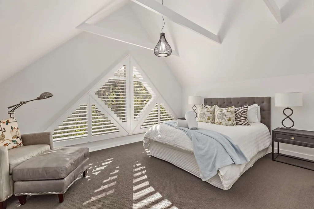 Kangaroo Valley Light-filled attic bedroom with triangular window, cozy bed, patterned pillows, armchair, and modern lamp.
