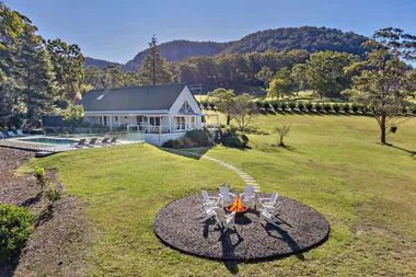 White cabin with a pool and fire pit in a grassy area, surrounded by trees and hills on a sunny day.