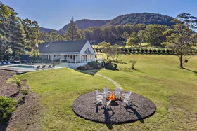 White cabin with a pool and fire pit in a grassy area, surrounded by trees and hills on a sunny day.