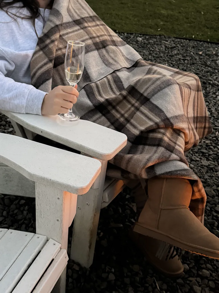 Person sitting outside wrapped in a plaid blanket, holding a champagne glass, wearing tan boots.