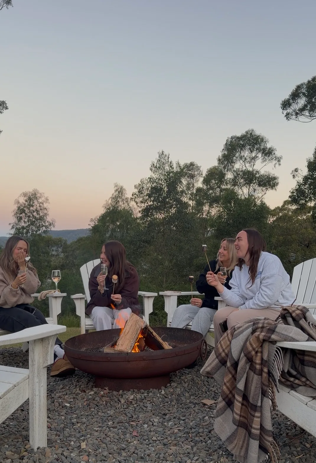 Four people sitting around a campfire at sunset, toasting marshmallows and holding wine glasses.