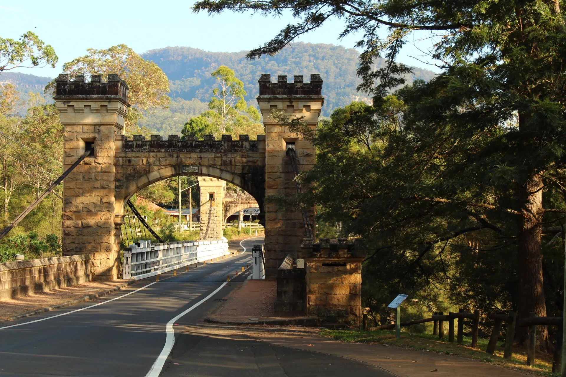 Historic sandstone suspension bridge with towers surrounded by lush greenery and mountains.