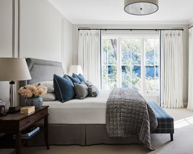 A light-filled bedroom with a cream coloured carpet, white walls, white curtains open to reveal a leafy backyard. A grey bed is in the centre of the room, with a grey quilt folded over the end of the white bed sheets and plenty of white and navy pillows on top. A bedside table is beside the bed, with a lamp, blue and white vase with peach toned dahlias inside and a wooden box.